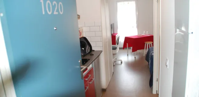 La porte d'entrée bleu ouverte sur le studio, on voit la petite kitchenette et la salle à manger.